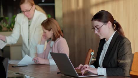 Nahaufnahme-Eines-Selbstbewussten-Brünetten-Mädchens-Mit-Runder-Brille-Und-Business-Kleidung,-Das-An-Einem-Laptop-Arbeitet-Und-Auf-Der-Tastatur-Tippt,-Während-Es-Zusammen-Mit-Seinen-Kollegen-Am-Tisch-Im-Büro-Arbeitet