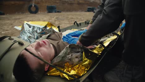 Close-up-providing-first-aid-to-wounded-soldiers-on-the-battlefield-under-the-supervision-of-a-military-medic.-Confident-male-medic-in-blue-rubber-medical-gloves-stops-the-blood-of-a-wounded-soldier-on-a-stretcher-in-a-combat-area