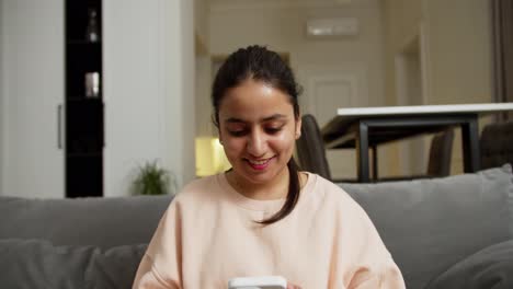 Happy-brunette-Indian-girl-in-light-pink-on-a-gray-sofa-during-her-relaxation-and-typing-on-social-networks-using-a-white-smartphone-at-home-in-a-modern-apartment