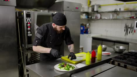 Confident-male-chef-in-a-black-uniform-with-a-beard-with-protective-rubber-gloves-puts-vegetables-on-a-plate-with-salad-and-twists-cucumber-slices-while-preparing-a-dish-in-the-kitchen-in-a-restaurant