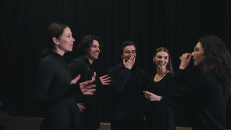 Un-Grupo-De-Estudiantes-Universitarios-De-Teatro-Felices-Y-Confiados,-Vestidos-Con-Trajes-Negros,-Se-Paran-En-El-Escenario-Cerca-De-Una-Cortina-Negra-Y-Se-Comunican-Expresivamente-Entre-Ellos.