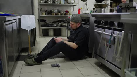 A-tired-male-chef-in-a-black-uniform-sits-on-the-floor-and-checks-social-networks-using-his-phone-during-a-short-break-in-the-middle-of-a-busy-working-day-in-the-kitchen-of-a-restaurant