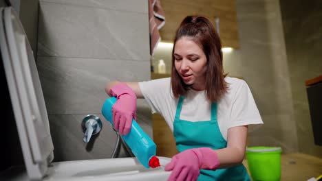 Una-Joven-Morena-Feliz,-Señora-De-La-Limpieza-Con-Una-Camiseta-Blanca-Y-Un-Delantal-Azul,-Vierte-Detergente-Azul-En-Las-Paredes-Del-Inodoro-Del-Baño-Durante-La-Limpieza