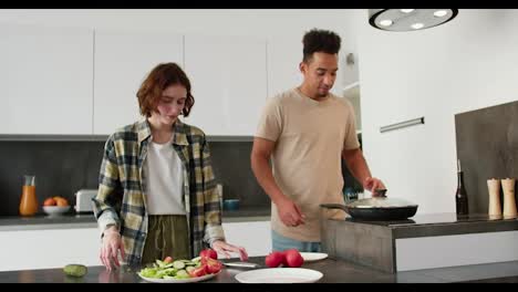 Portrait-of-a-young-Black-skinned-brunette-man-in-a-cream-t-shirt-and-his-young-adult-girlfriend-with-a-bob-hairstyle-they-present-their-breakfast-of-scrambled-eggs-and-fresh-salad-during-their-breakfast-preparation-in-a-modern-kitchen