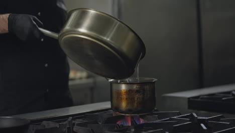 Un-Cocinero-Con-Uniforme-Negro-Vierte-Agua-De-Una-Cacerola-Grande-En-Una-Cacerola-Pequeña-Sobre-Un-Quemador-De-Gas-En-La-Cocina-De-Un-Restaurante.