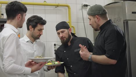 Two-chefs-in-black-uniforms-together-with-waiters-in-white-shirts-communicate-about-the-prepared-dishes-and-solve-their-problems-while-working-in-the-kitchen-and-restaurant.-Cooks-and-waiters-solve-problems-and-questions-that-arise-regarding-dishes-and-their-quality-in-the-kitchen