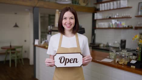 Portrait-of-a-happy-brunette-waitress-girl-in-a-yellow-apron-holding-in-her-hands-a-sign-with-an-open-sign-in-a-cafe