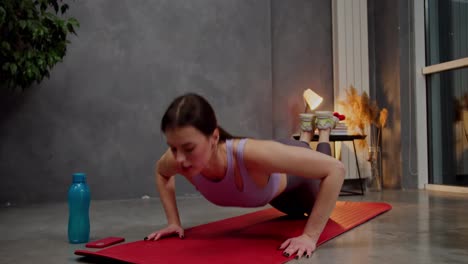 Confident-athletic-brunette-girl-in-a-purple-top-and-gray-sweatpants-on-a-red-sports-mat-does-sports-runs-in-place-and-does-push-ups-in-a-modern-apartment-near-a-large-indoor-plant