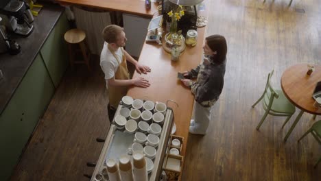 Top-view-of-a-brunette-girl-in-a-gray-sweater-makes-an-order-at-the-counter-from-the-barista-of-a-blond-guy-in-a-cafe