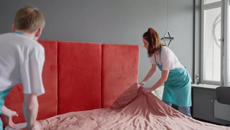 A-brunette-cleaning-lady-in-a-white-T-shirt-and-a-blue-apron-together-with-her-blond-male-colleague-makes-the-bed-with-a-pink-blanket-and-puts-gray-pillows-while-cleaning-the-bedroom