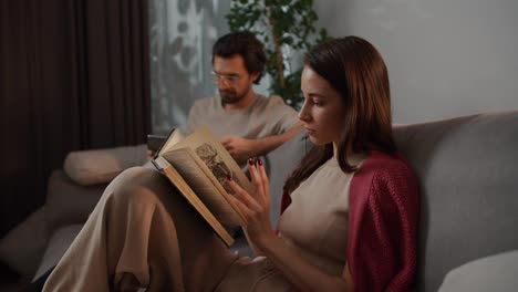 Side-view-of-a-young-brunette-girl-in-a-red-sweater-reading-a-book-while-sitting-on-a-gray-sofa-with-her-boyfriend-in-glasses-who-is-looking-at-a-tablet-in-a-modern-apartment