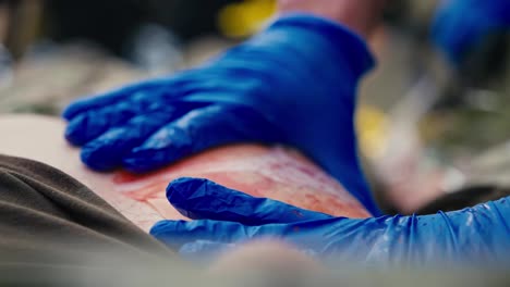 Close-up-a-confident-medic-in-blue-rubber-gloves-applies-a-special-large-plaster-to-the-wound-site-of-a-soldier-to-avoid-blood-loss-in-combat-conditions-during-first-aid