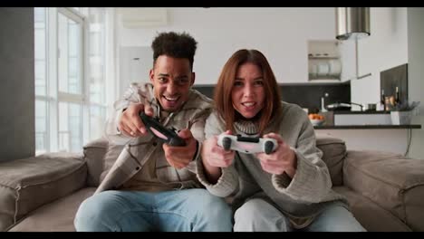 A-happy-Black-skinned-man-with-a-brunette-and-his-girlfriend-in-a-sweater-play-a-video-game-using-a-joystick.-Then-the-man-wins-and-the-girl-is-sad-about-her-defeat-in-a-modern-apartment-Sitting-on-the-sofa