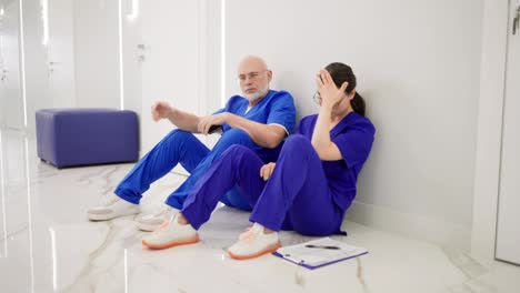 A-confident-and-tired-doctor-an-elderly-man-with-glasses-with-a-white-beard-in-a-blue-uniform-sits-with-his-colleague-a-brunette-girl-on-the-floor-after-a-hard-day-at-work-during-a-break-between-patients-in-a-modern-clinic