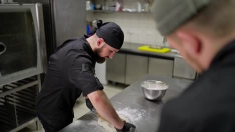 Professional-male-chef-in-black-uniform-with-bandana-rolling-out-dough-in-kitchen-using-dough-and-bowls.-Confident-male-chef-rolls-out-dough-using-a-rolling-pin-and-tells-a-trainee-how-to-roll-it-out-correctly