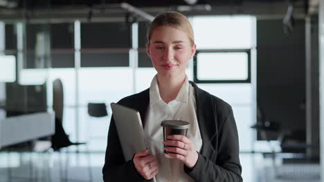 Porträt-Eines-Glücklichen-Blonden-Mädchens-Im-Business-Anzug,-Das-Eine-Tasse-Kaffee-Und-Einen-Laptop-In-Den-Händen-Hält,-Während-Es-In-Einem-Modernen-Büro-Arbeitet