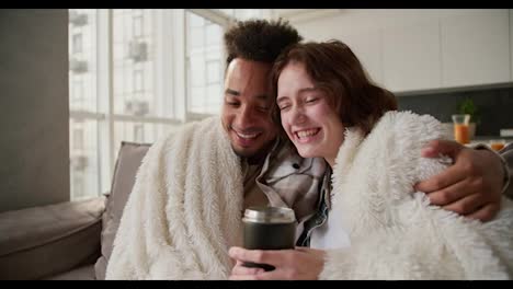 A-happy-young-man-with-Black-skin-color-in-a-checkered-cream-shirt-sits-on-a-modern-sofa-wrapped-in-a-white-woolen-blanket-with-his-young-girlfriend-who-drinks-tea-from-a-black-glass-they-hug-and-spend-time-together-in-a-modern-apartment