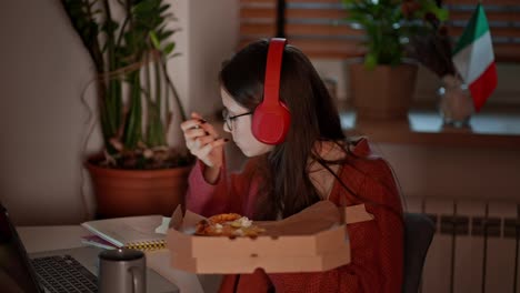 Side-view-of-a-confident-brunette-girl-in-red-wireless-headphones-studying-foreign-languages-near-the-flag-of-Italy-using-online-lessons-on-a-laptop-and-taking-notes-in-her-notes-while-eating-pizza-in-a-modern-apartment-in-the-kitchen