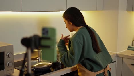 A-happy-brunette-girl-with-Brown-skin-in-a-green-jacket-and-a-brown-apron-broadcasts-and-broadcasts-her-culinary-blog-while-cooking-in-a-gray-shiny-pan-in-a-modern-kitchen