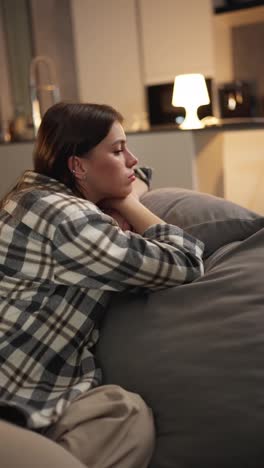 Vertical-video-of-a-tired-and-bored-brunette-girl-in-a-checkered-shirt-lies-on-the-sofa-leaning-on-the-back-and-on-the-pillow-in-a-modern-apartment-at-home-in-the-evening