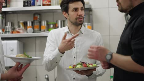 Un-Camarero-Con-Camisa-Blanca-Se-Comunica-Sobre-Los-Platos-Preparados-Por-El-Chef-Con-El-Autor-De-Platos-Y-Ensaladas-Presentando-Sus-Quejas-Y-Preguntas-En-La-Cocina-De-Un-Restaurante.