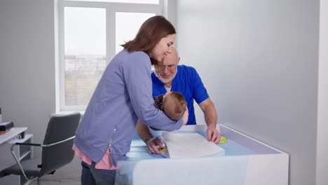 Confident-brunette-girl-puts-her-baby-girl-on-the-bed-in-front-of-an-experienced-male-pediatrician-doctor-in-a-blue-uniform-in-a-pediatric-doctors-office-in-a-modern-clinic