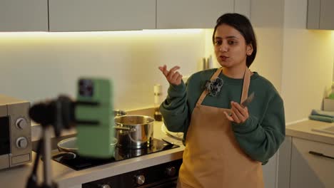 Una-Chica-Morena-Feliz-Con-Una-Chaqueta-Verde-Y-Un-Delantal-Beige-Administra-Su-Blog-Culinario-Y-Usa-Su-Teléfono-Para-Contar-Cómo-Preparar-Adecuadamente-La-Comida-Y-Otros-Secretos-Culinarios-En-Una-Cocina-Moderna.