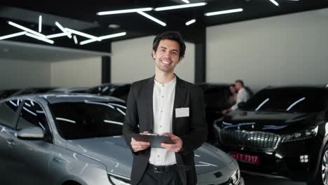 Porträt-Eines-Selbstbewussten-Und-Glücklichen-Jungen-Brünetten-Mannes-In-Einem-Business-Anzug.-Assistent-In-Einem-Modernen-Autohaus.-Ein-Selbstbewusster-Brünetter-Mann-In-Einem-Business-Anzug-Und-Einem-Weißen-Hemd-Steht-Mitten-In-Einem-Autohaus-Mit-Einem-Tablet-In-Den-Händen