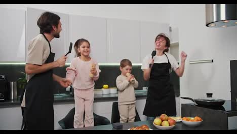 Familia-Feliz,-Hombre-Moreno-Con-Barba-Incipiente-Con-Delantal-Negro-Junto-Con-Su-Esposa-Morena-Y-Dos-Niños-Pequeños-Bailando-Y-Divirtiéndose-En-Una-Cocina-Moderna-Antes-De-Comenzar-El-Desayuno-En-Un-Apartamento-Moderno-Por-La-Mañana