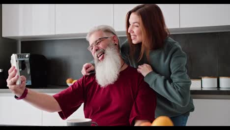 A-happy-elderly-man-with-gray-hair-and-a-lush-beard-in-a-red-T-shirt-takes-a-selfie-using-a-White-phone-with-his-adult-brunette-daughter-in-a-green-sweater-in-a-modern-apartment