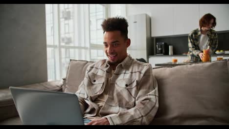 Happy-Black-skinned-brunette-man-with-stubble-in-a-checkered-cream-shirt-works-with-his-gray-laptop-and-is-very-happy-about-his-success-while-working-from-home-in-a-modern-studio-apartment