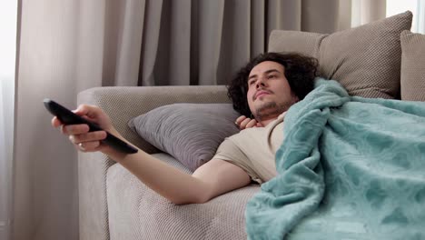 Confident-guy-brunette-with-curly-hair-lying-on-the-sofa-covered-himself-in-front-of-him-and-rewinding-channels-using-the-remote-control-at-home