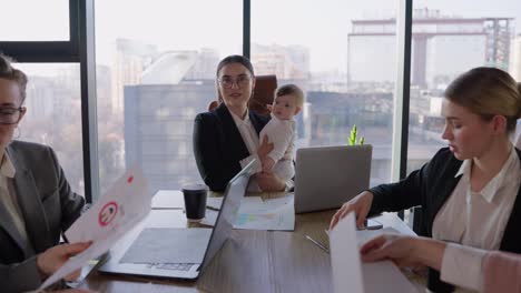 Portrait-of-a-confident-brunette-girl-in-round-glasses-in-a-business-suit-who-sits-at-a-table-in-the-office-and-holds-a-small-infant-child-in-her-arms-during-a-meeting-and-thinking-about-her-idea-and-plan-in-an-office-with-large-windows-at-the-table