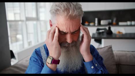 Close-up-of-an-elderly-man-with-gray-hair-and-a-full-beard-in-a-blue-shirt-kneads-and-massages-his-temples-during-a-headache-in-a-modern-apartment