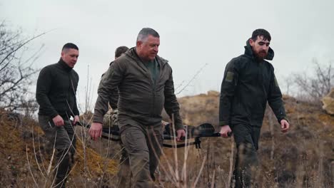 Four-confident-male-experienced-soldiers-carry-a-wounded-soldier-in-a-camouflage-uniform-and-a-brunette-man-in-dark-green-clothes-stumbles-dropping-a-wounded-soldier-during-fighting-in-the-steppe