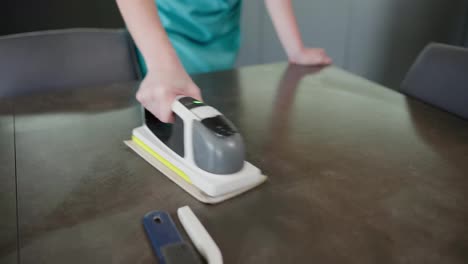 Close-up-a-girl-in-a-blue-apron-polishes-the-surface-of-a-glossy-table-in-the-kitchen-using-a-device-for-cleaning-an-apartment-and-kitchen