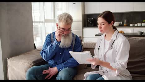 An-elderly-man-with-gray-hair-and-a-lush-beard-puts-on-glasses-to-read-what-a-brunette-woman-doctor-in-a-white-coat-wants-to-convey-to-him-during-a-home-examination-on-the-sofa-in-a-modern-apartment