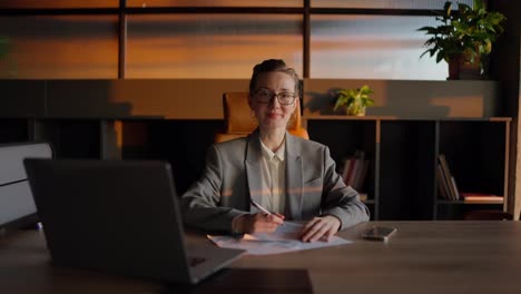 Portrait-of-a-confident-middle-aged-blonde-businesswoman-with-glasses-in-a-gray-jacket-who-sits-in-front-of-sheets-of-papers-with-a-writing-pen-in-her-hands-at-a-wooden-table-in-a-sunny-office