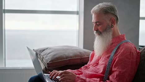 Side-view-of-an-elderly-man-with-gray-hair-and-a-full-beard-with-a-ring-in-his-ear-and-a-pink-shirt-sitting-on-a-modern-sofa-and-typing-on-his-gray-laptop-in-a-modern-apartment-overlooking-the-sea