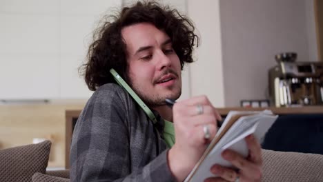 Close-up-focused-happy-brunette-guy-with-curly-hair-and-mustache-in-a-checkered-shirt-listens-communicates-on-the-phone-and-writes-down-important-points-in-a-notepad-during-his-working-day-at-home