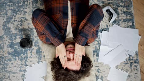 Camera-rotation-Tired-brunette-guy-with-curly-hair-lies-on-the-floor-on-a-pillow-around-him-sheets-of-paper-coffee-laptop-and-smartphone-at-home-in-a-modern-apartment