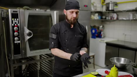 Confident-male-chef-with-a-beard-in-a-black-uniform-puts-on-gloves-and-starts-preparing-a-salad-in-a-modern-kitchen-in-a-restaurant.-Start-of-the-working-day-in-the-kitchen-and-use-of-protective-gloves