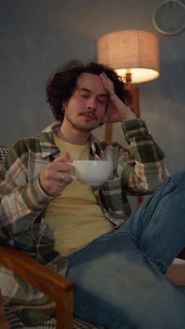 Vertical-video-of-a-tired-brunette-guy-in-a-checkered-shirt-drinking-a-hot-drink-from-a-cup-and-thinking-about-his-life-near-a-lamp-at-home-in-the-evening