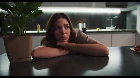 A-sad,-thoughtful-brunette-girl-in-a-beige-T-shirt-leans-on-the-table-and-thinks-about-something-sad-near-a-houseplant-on-a-black-table-in-the-kitchen-in-a-modern-apartment