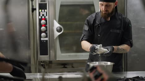 Confident-professional-chef-in-a-black-uniform-with-a-beard-places-dough-on-the-table-with-flour-and-rolls-it-out-with-his-hands-while-preparing-pizza-in-the-kitchen-in-a-restaurant