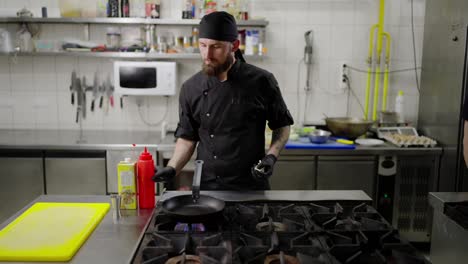 Confident-male-chef-in-black-protective-clothing-with-a-beard-and-bandana-pours-oil-into-a-frying-pan-on-the-stove-while-starting-to-prepare-a-dish-in-a-modern-kitchen-in-a-restaurant