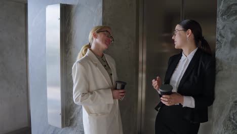 Couple-of-happy-businesswomen-in-a-white-and-black-business-suit-talking-while-drinking-coffee-during-a-break-from-work-in-a-modern-office