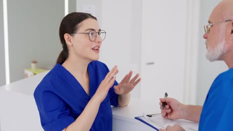 Una-Chica-Morena-Segura-Y-Feliz-Con-Gafas-Redondas-Y-Un-Uniforme-Azul-Habla-Con-Su-Colega,-Un-Médico-Experimentado-Con-Barba-Blanca,-Sobre-La-Jornada-Laboral-En-Una-Clínica-Moderna-Y-Luminosa.