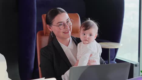 Close-up-of-a-confident-brunette-businesswoman-girl-in-round-glasses-and-in-business-clothes-sitting-at-the-office-desk-and-holding-her-small-baby-child-in-her-arms-during-a-business-meeting-in-the-office