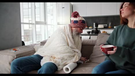 A-brunette-girl-in-a-green-jacket-brings-a-mug-of-tea-to-her-sick-father-of-old-age-with-a-thick-gray-beard-in-a-white-plaid-and-takes-care-of-him-during-the-mans-illness-in-a-modern-apartment